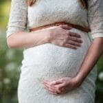 A pregnant woman in a lace dress gently cradling her belly in an outdoor setting.