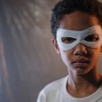 Portrait of a young child wearing a white eye mask in an indoor setting with dramatic lighting.