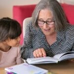 A joyful grandmother reading a book with her granddaughter indoors, bonding over learning.