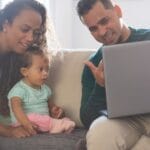 A cheerful family of three enjoying quality time together on their living room sofa, engaging with a laptop.