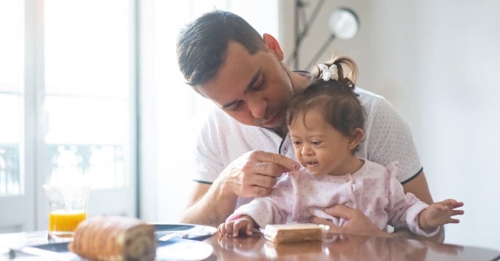 Father lovingly feeds daughter with Down syndrome during breakfast at home.