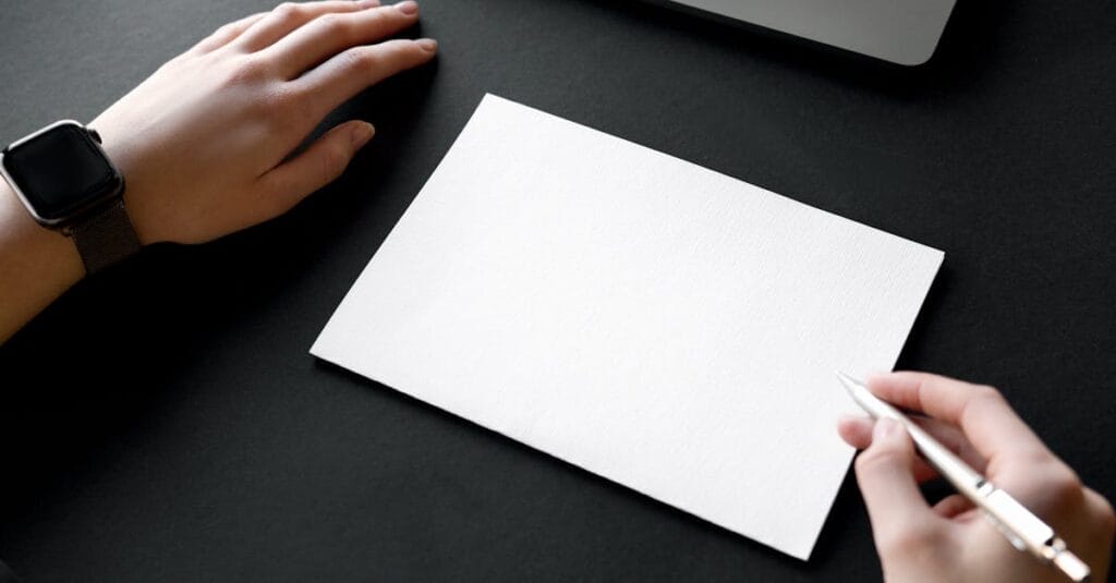 Hands positioned over blank paper ready to write with a pen, accompanied by a laptop.
