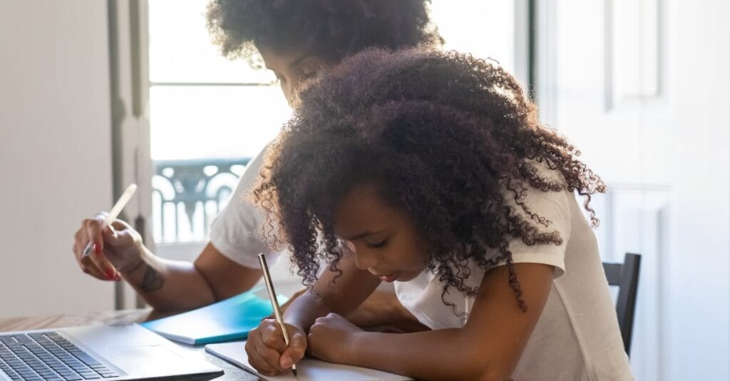 Young girl with afro hair studying with parent at home, focused on writing tasks.