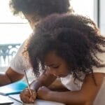 Young girl with afro hair studying with parent at home, focused on writing tasks.