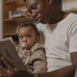 A father reads a book to his baby while sitting on a couch, creating a warm family moment.