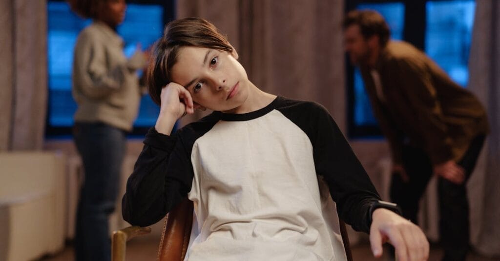 A young boy in a black and white shirt looks sad as his parents argue in the background.