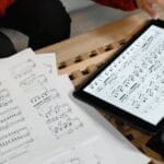 A person reviewing and composing sheet music using both paper and digital formats on a table.