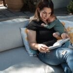 A serene moment of a mother reading while holding her sleeping baby on an outdoor sofa.