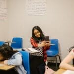 A diverse group of students in a classroom setting, one using a smartphone.