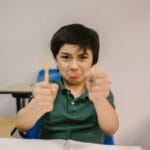 A young boy in a classroom showing frustration, holding broken pencils.