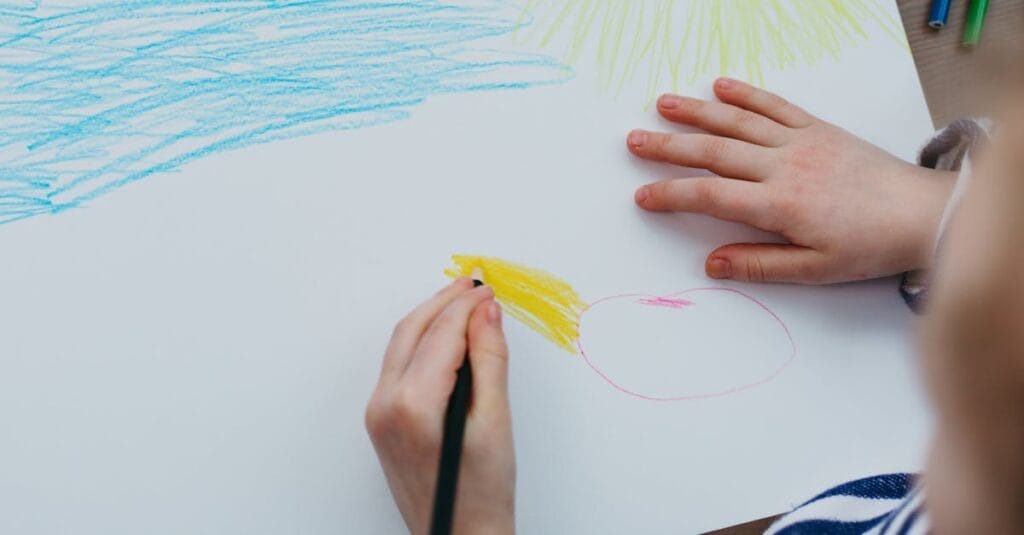 A child drawing a colorful sun and sky with crayons on a white sheet of paper.