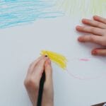 A child drawing a colorful sun and sky with crayons on a white sheet of paper.