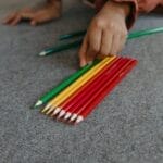 A hand organizes vibrant colored pencils on a carpeted floor.