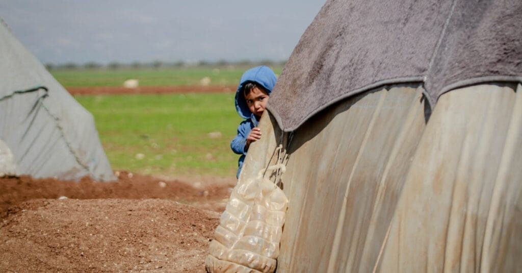 A young boy in a blue hoodie peeking from behind a tent in a refugee camp in Idlib, Syria.