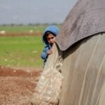 A young boy in a blue hoodie peeking from behind a tent in a refugee camp in Idlib, Syria.