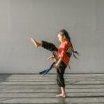 A young girl practicing a martial arts kick in a sunlit dojo indoor environment.