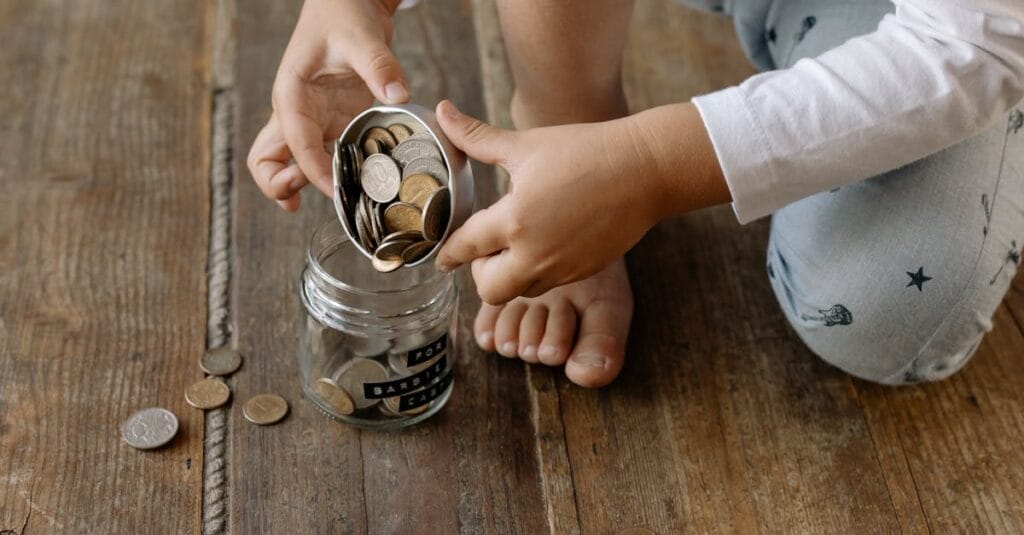 A child adds coins into a glass jar labeled for savings on a wooden floor.