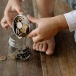 A child adds coins into a glass jar labeled for savings on a wooden floor.