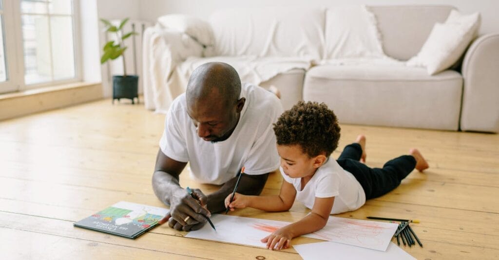 Father and son spend quality time drawing together on a wooden floor at home.