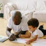 Father and son spend quality time drawing together on a wooden floor at home.