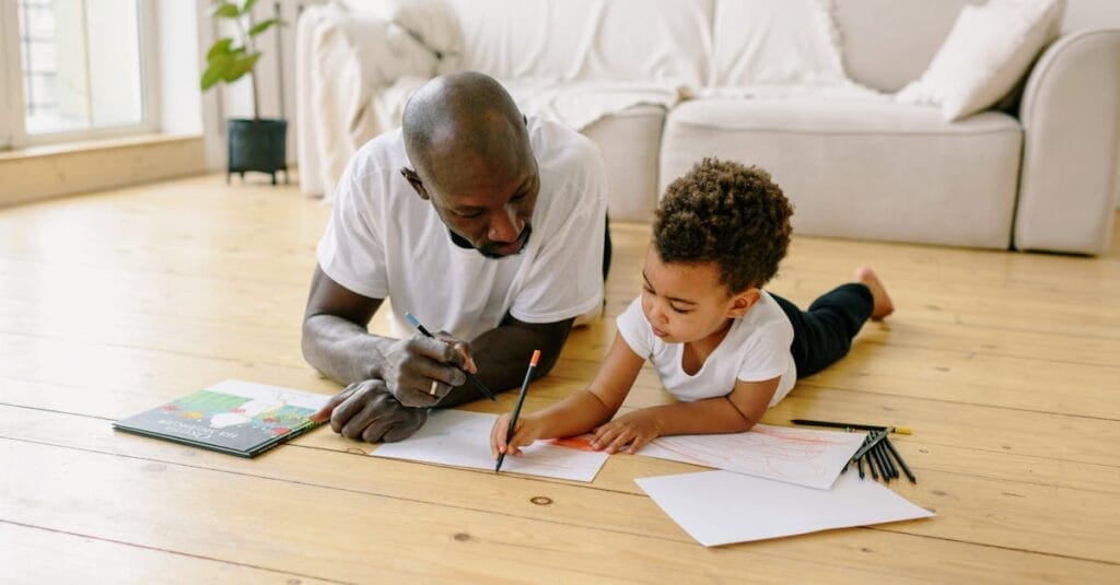 A father and son bond while drawing with colored pencils on the floor indoors.