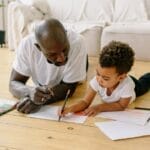 A father and son bond while drawing with colored pencils on the floor indoors.