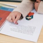 Child's hand pointing at illustrations in a colorful book on a wooden table.