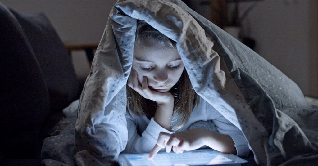 A child using a tablet under a blanket in a dark room, creating a cozy bedtime atmosphere.