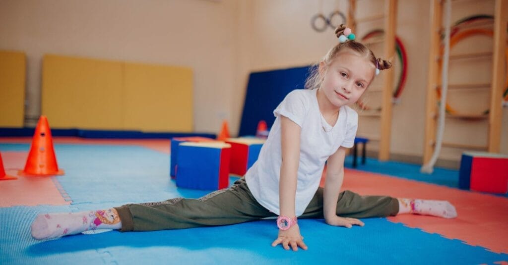 Young girl showing flexibility while practicing splits indoors on colorful mats in a playroom setting.