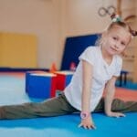 Young girl showing flexibility while practicing splits indoors on colorful mats in a playroom setting.