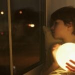 A young boy holding a glowing orb, looking out a window at night.