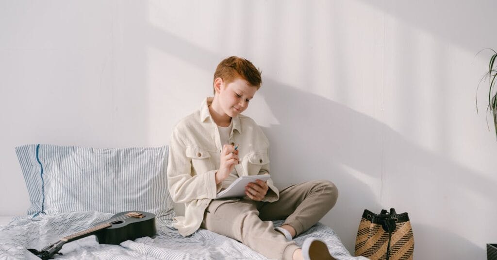 Young boy writing in a notebook while sitting on a bed with a guitar nearby in a bright room.
