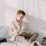 Young boy writing in a notebook while sitting on a bed with a guitar nearby in a bright room.