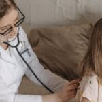 A doctor with a stethoscope examining a smiling girl during a medical check-up at home.