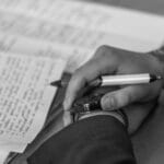 Close-up of a hand holding a pen over pages of writing on a desk.