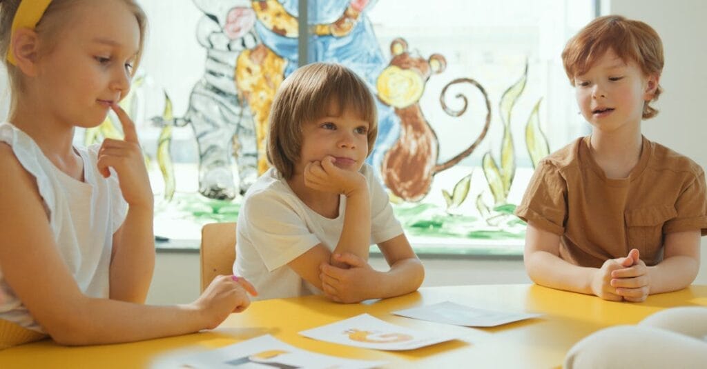 Three children sitting at a yellow table in a classroom, engaging in a learning activity.