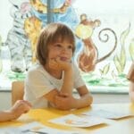 Three children sitting at a yellow table in a classroom, engaging in a learning activity.