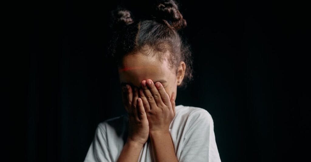 A child covers their face in a studio portrait with a dark background, creating a mysterious and innocent vibe.