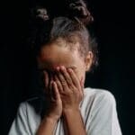 A child covers their face in a studio portrait with a dark background, creating a mysterious and innocent vibe.