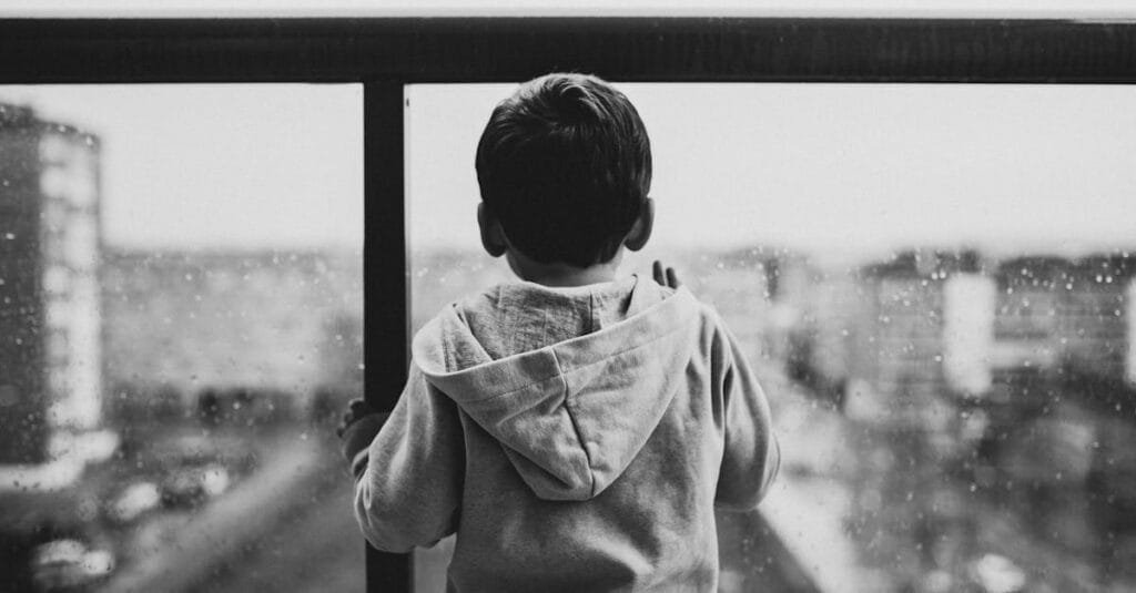 A young boy in a hoodie looks out a rainy window, creating a moody black-and-white scene.