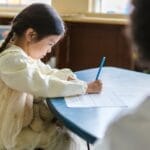 A young child intensely focused on writing at a classroom table, showing concentration and learning.