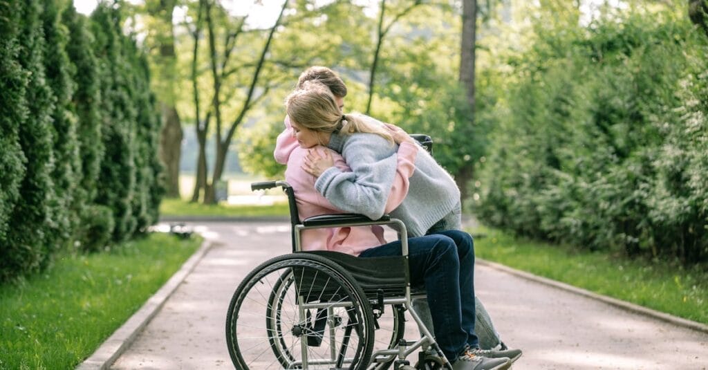 A couple lovingly embraces on a sunny day in a park, with one using a wheelchair.