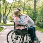 A couple lovingly embraces on a sunny day in a park, with one using a wheelchair.