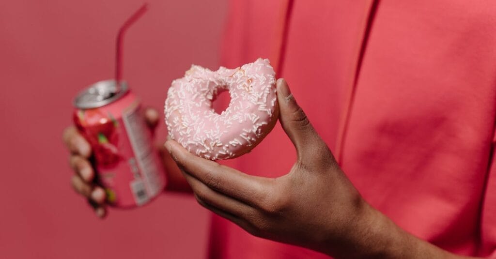 Close-up of a hand holding a pink doughnut and a soda can against a pink backdrop.