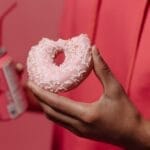 Close-up of a hand holding a pink doughnut and a soda can against a pink backdrop.