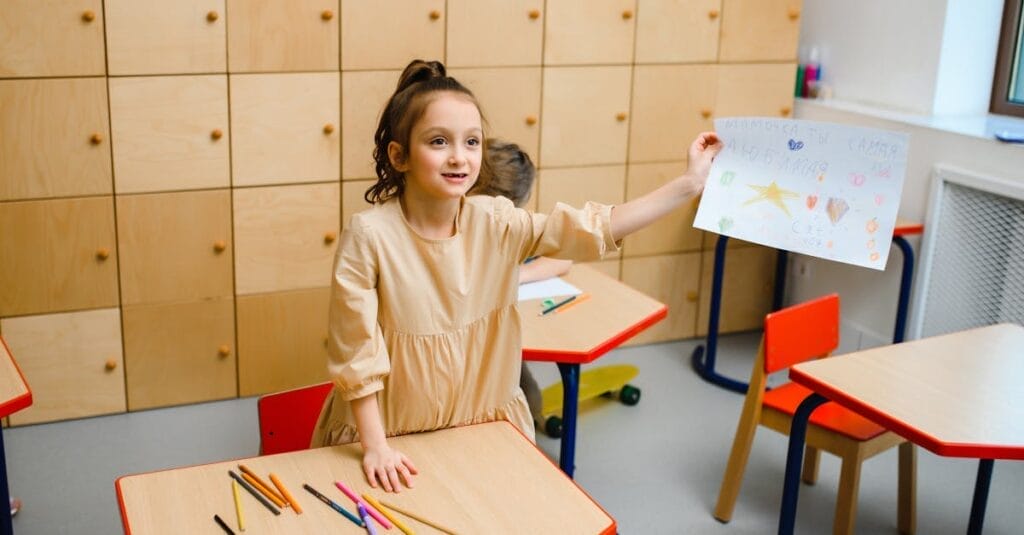 Smiling girl proudly showing her drawing in a vibrant kindergarten classroom.