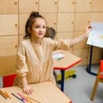 Smiling girl proudly showing her drawing in a vibrant kindergarten classroom.