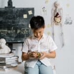 Asian boy solving Rubik's Cube in a classroom, with books and educational materials around.