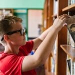 A young boy wearing glasses selects a book from a library bookshelf, emphasizing education and reading.