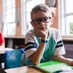Young boy with glasses in a classroom, pen in hand, looking thoughtful.